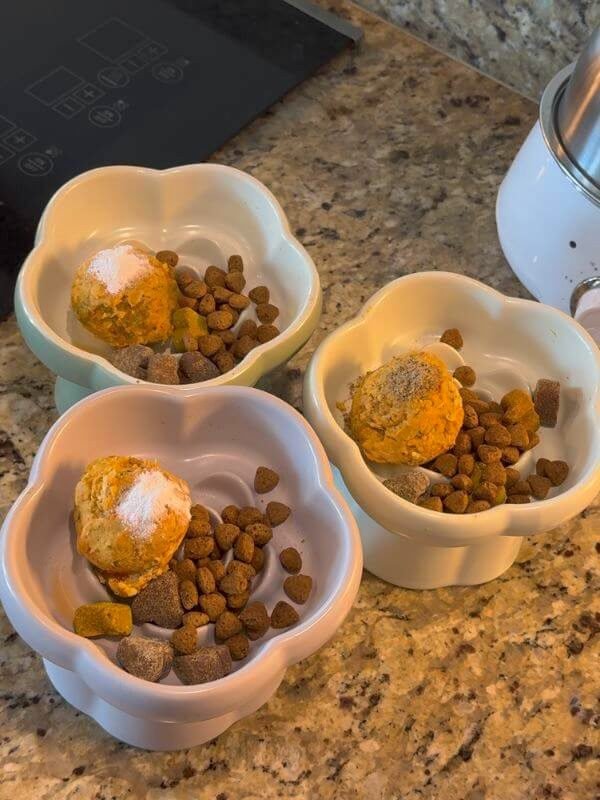 Three ceramic dog food bowls prepped with thawed homemade food scoops, prescription kibble, and daily supplements