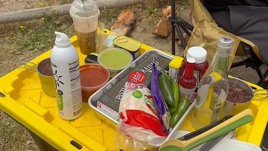 Everything we used for cooking every meal on a campfire grate laid out on a yellow camp table, including tortillas, salsa, peppers, seasonings and an iced coffee