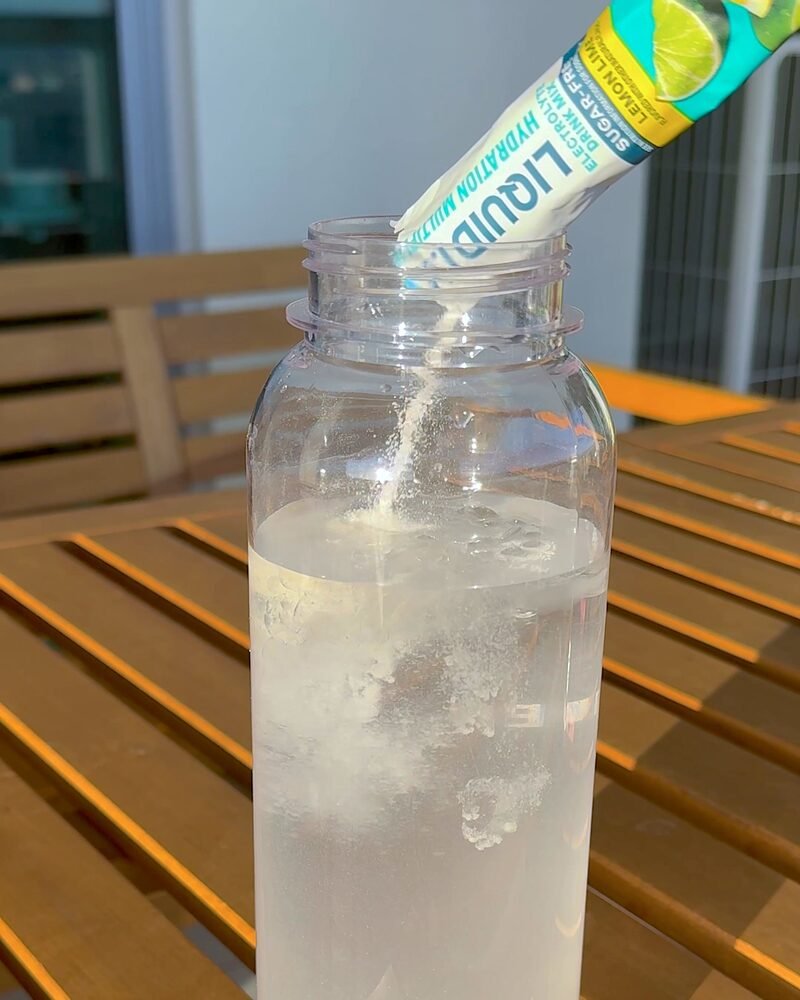 Electrolyte powder pouring from a stick pack into a clear bottle of ice water on a sunlit table