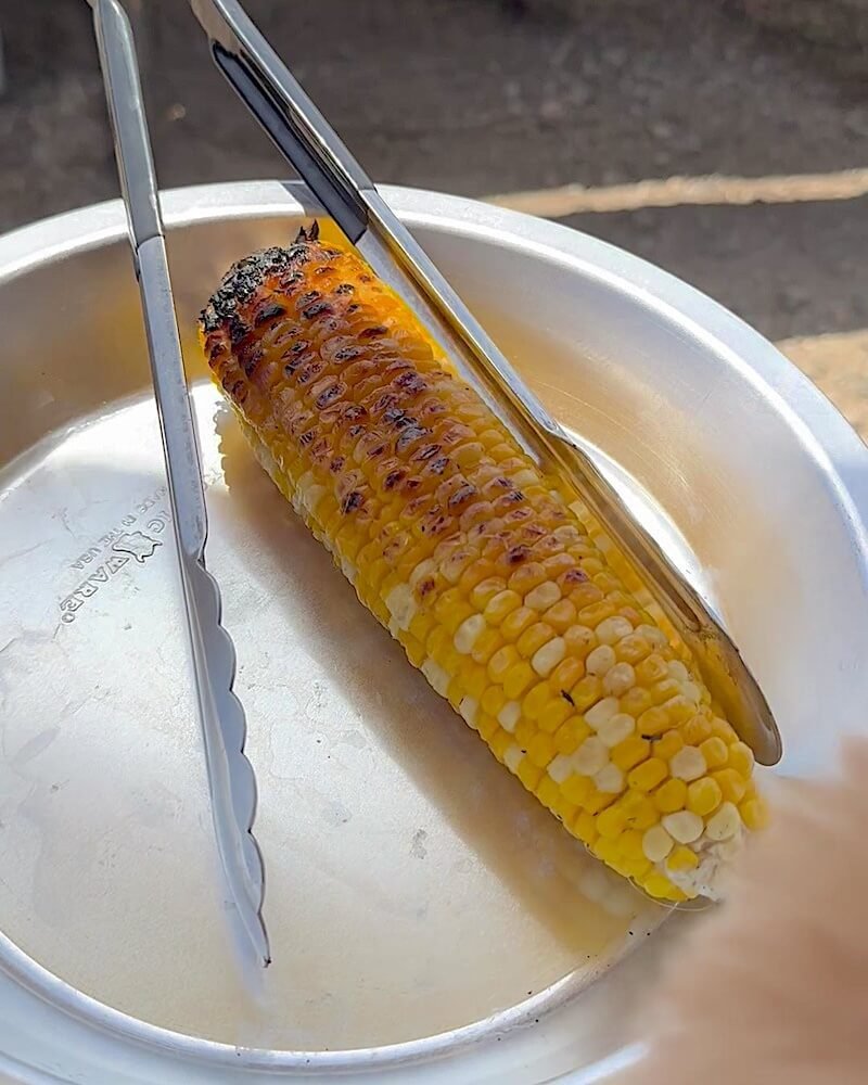 Corn on the cob charred in spots resting on a metal plate with a pair of tongs after cooking on a campfire grate