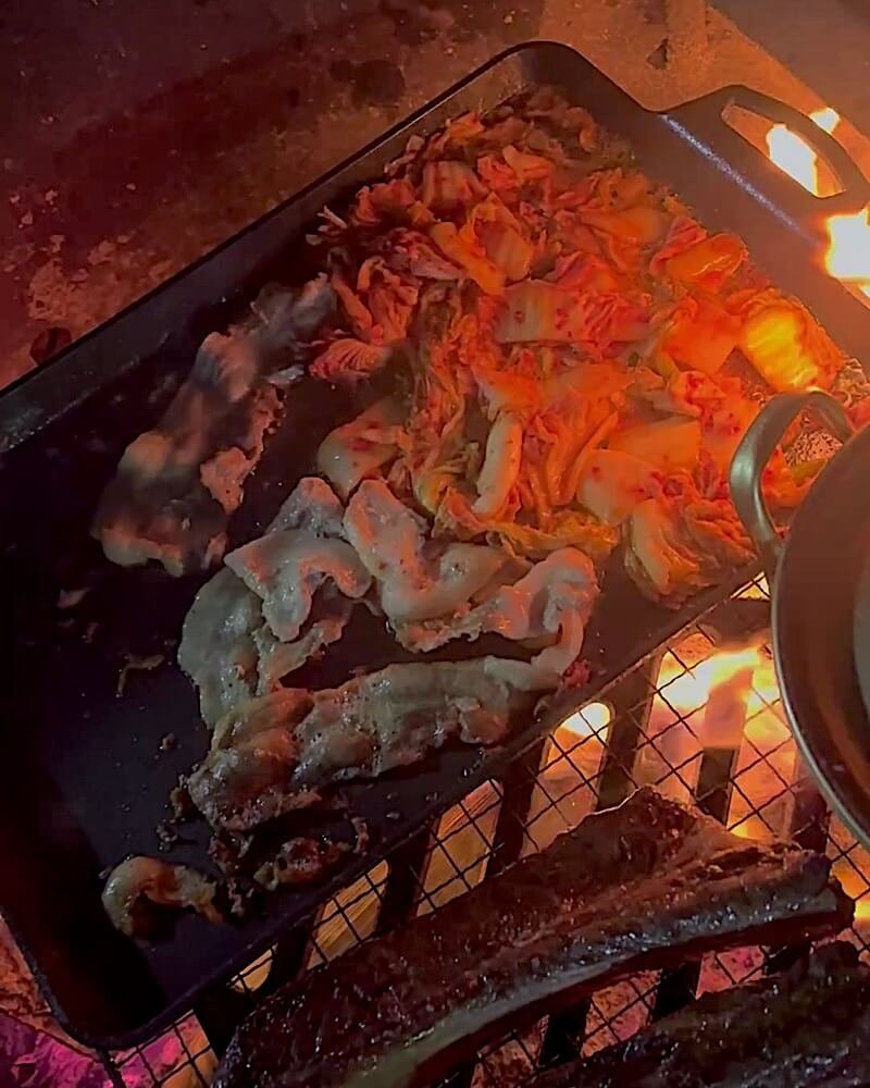Pork belly and kimchi cooking side by side on a flat cast iron pan set over a campfire grate with a steak on the grate below