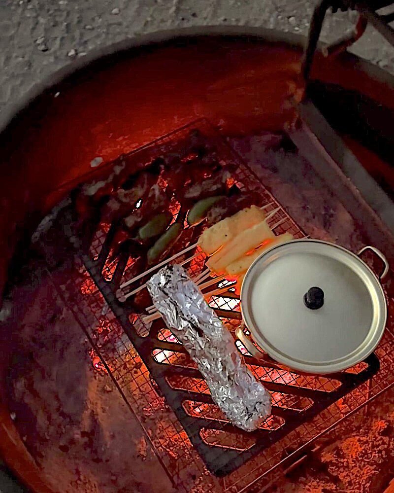 Small metal pot with a lid sitting on a campsite fire ring grate above a bed of glowing orange coals at night