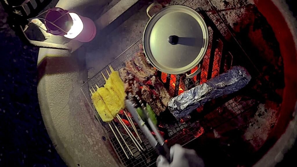 Overhead view of a campfire grate holding a covered pot, skewers, corn and a foil packet over glowing coals