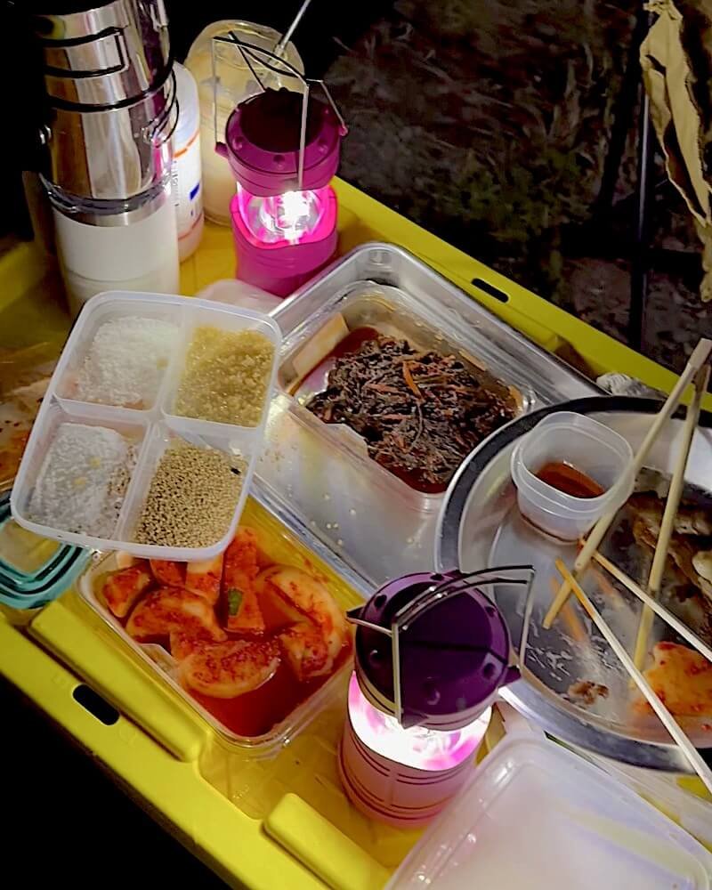 Food portioned into clear containers on a yellow camp table at night, lit by a small lantern, ready to be heated on the fire