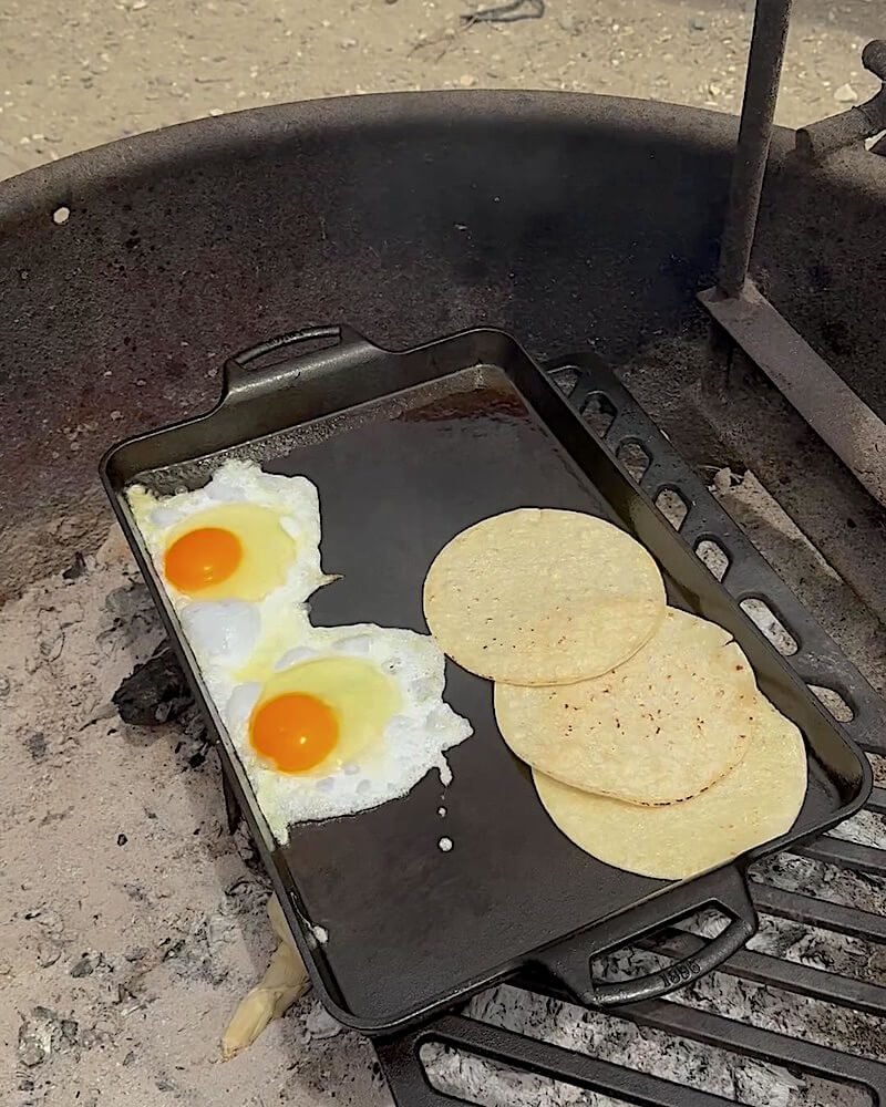 Two fried eggs and warming tortillas on a cast iron flat pan set on a campsite fire ring grate