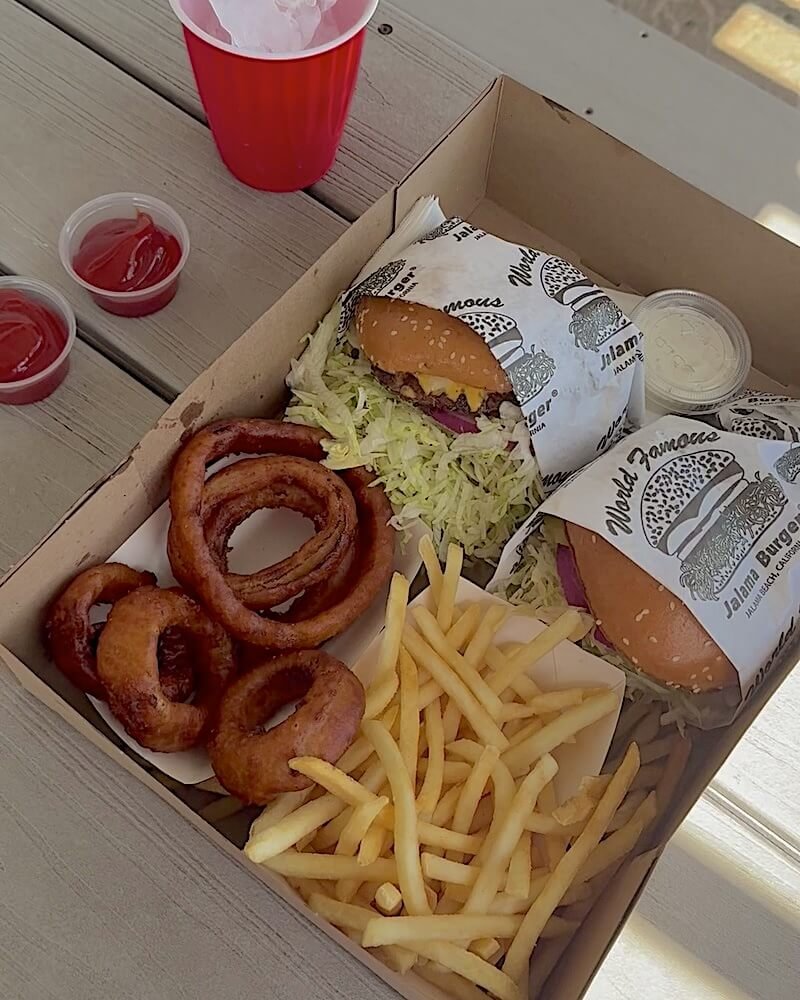 Two Jalama Burgers wrapped in paper with onion rings, fries and dipping sauces in a cardboard takeout box