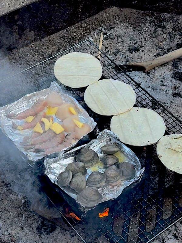 Clams in foil tray and steaks sizzling on campfire grill grate by the ocean