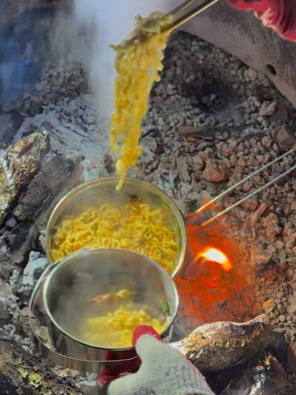 Hot steaming ramen cooking in metal pot over glowing campfire embers at night