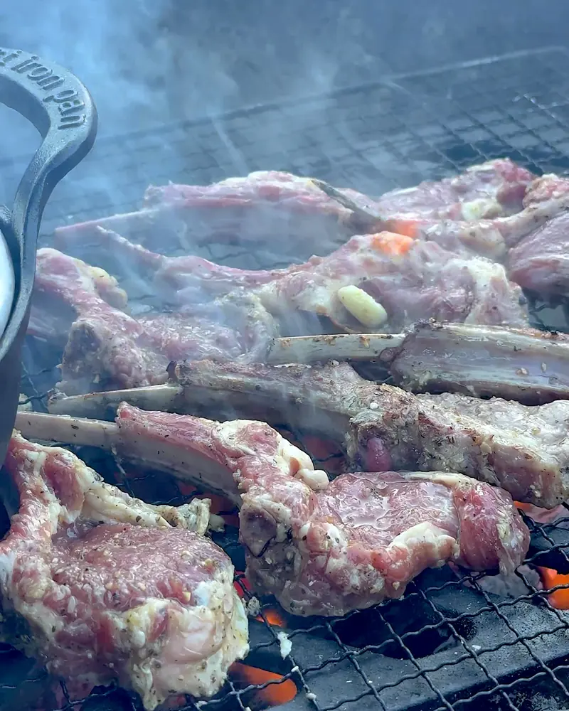 Cooking on a Fire Pit at Camp (6 Things We Made, From Lamb Chops to Pork Belly) Cooking on a fire pit: marinated lamb chops sizzling on a campfire grate with a cast iron griddle at the edge