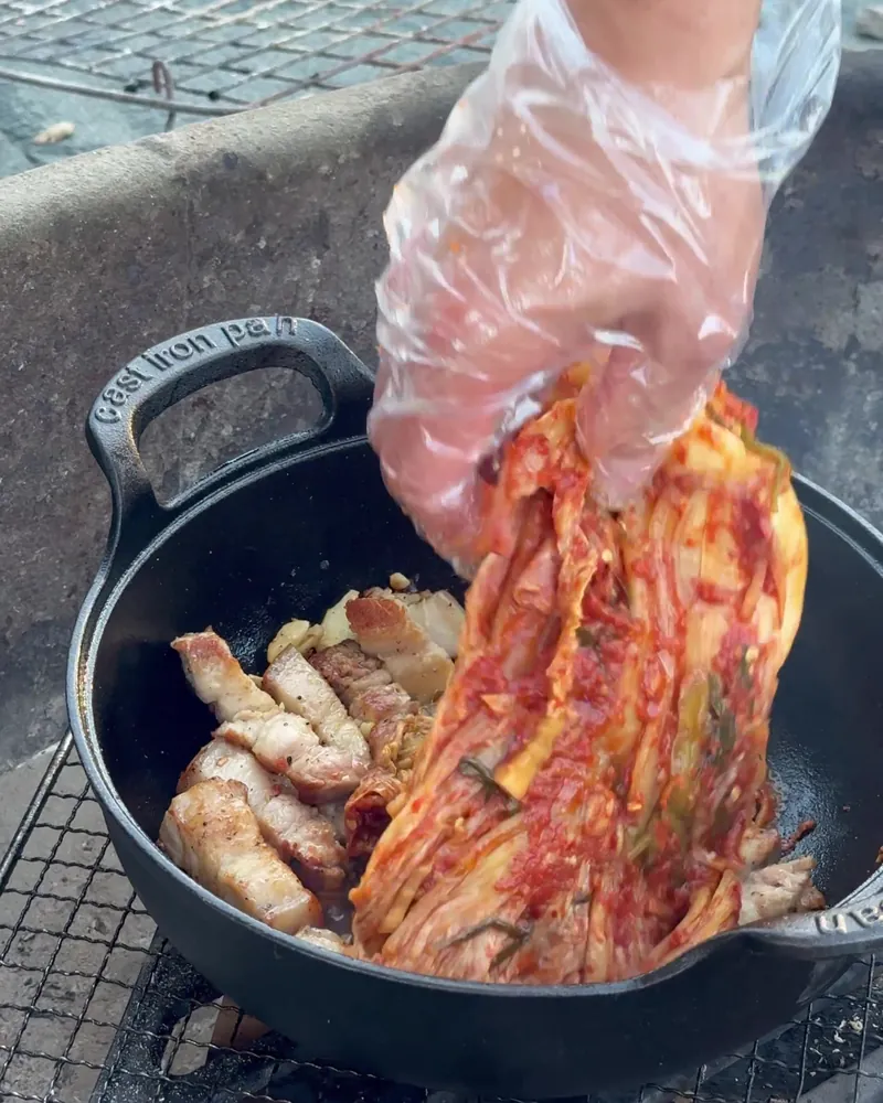 Camping Food List: What We Actually Ate in 3 Nights (Korean Dinners, Drinks, and the Dogs' Food) Hand in a disposable glove adding aged kimchi on top of browned pork belly in a cast iron pot over a campfire grate