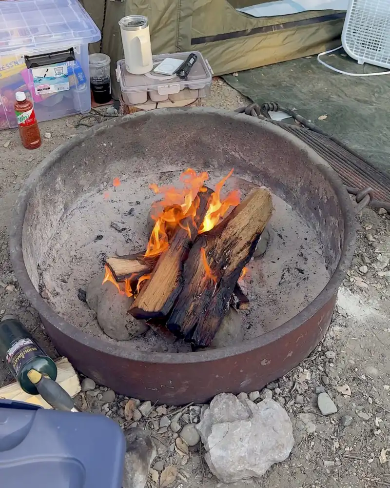 Cooking on a Fire Pit at Camp (6 Things We Made, From Lamb Chops to Pork Belly) Split firewood burning in the steel fire ring at our campsite, with a cooler and camp supplies around it