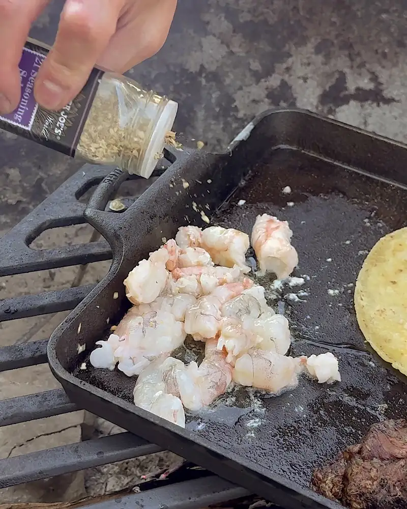 Easy Camping Breakfast: 4 Campfire Griddle Meals We Made (Nothing Prepped at Home) Hand shaking seasoning over thawed shrimp cooking on a cast iron griddle