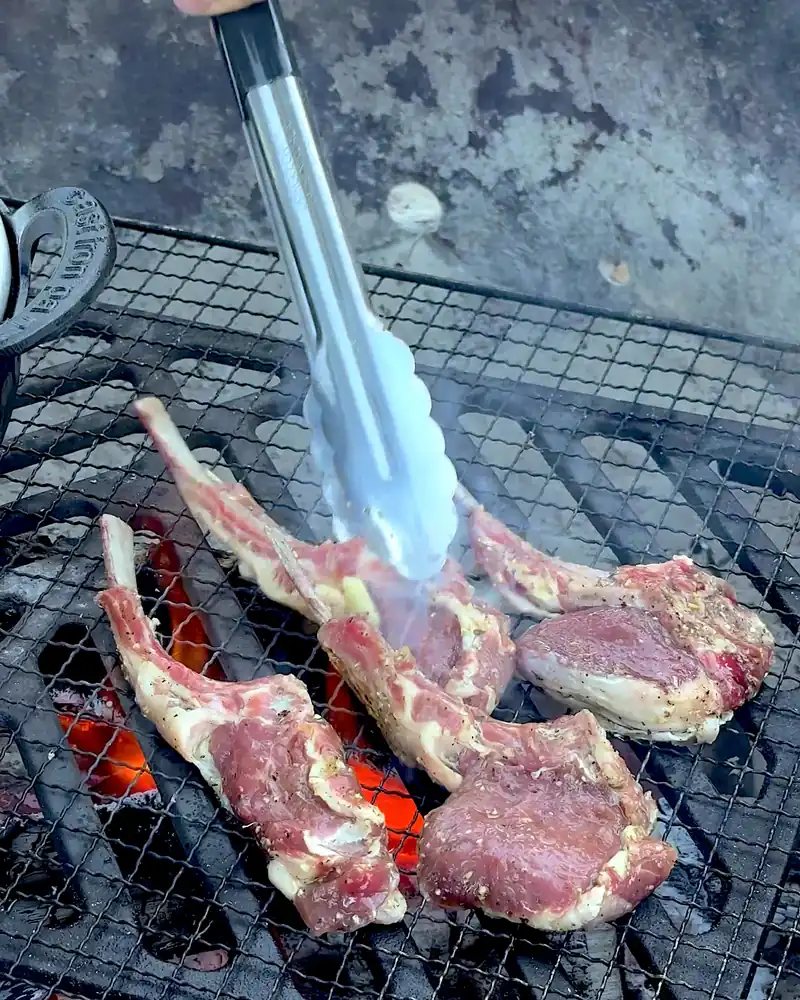 Cooking on a Fire Pit at Camp (6 Things We Made, From Lamb Chops to Pork Belly) Flipping lamb chops with long metal tongs over glowing coals on a campfire grate
