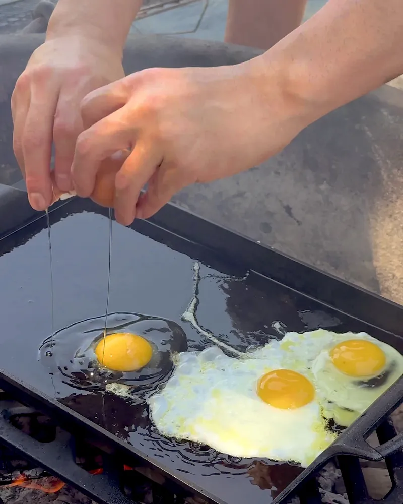 Easy Camping Breakfast: 4 Campfire Griddle Meals We Made (Nothing Prepped at Home) Hands cracking eggs onto a hot cast iron griddle over the fire