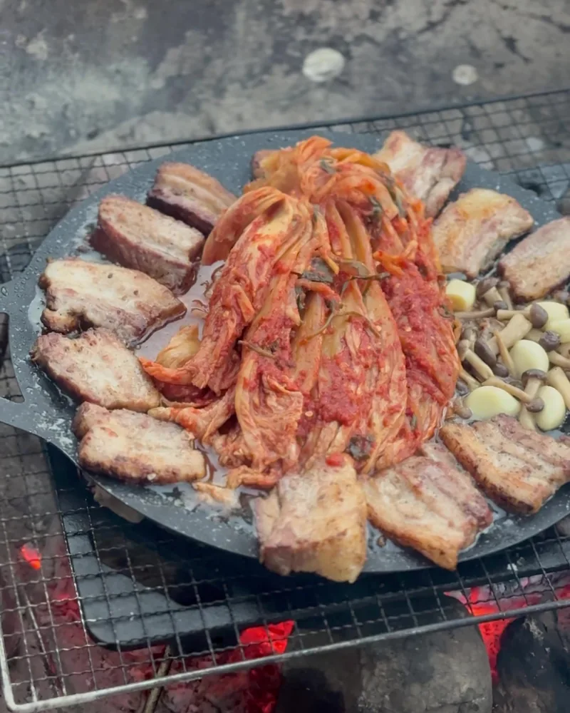 Camping Food List: What We Actually Ate in 3 Nights (Korean Dinners, Drinks, and the Dogs' Food) Sliced pork belly with a pile of kimchi, small mushrooms and garlic on a round griddle over campfire coals