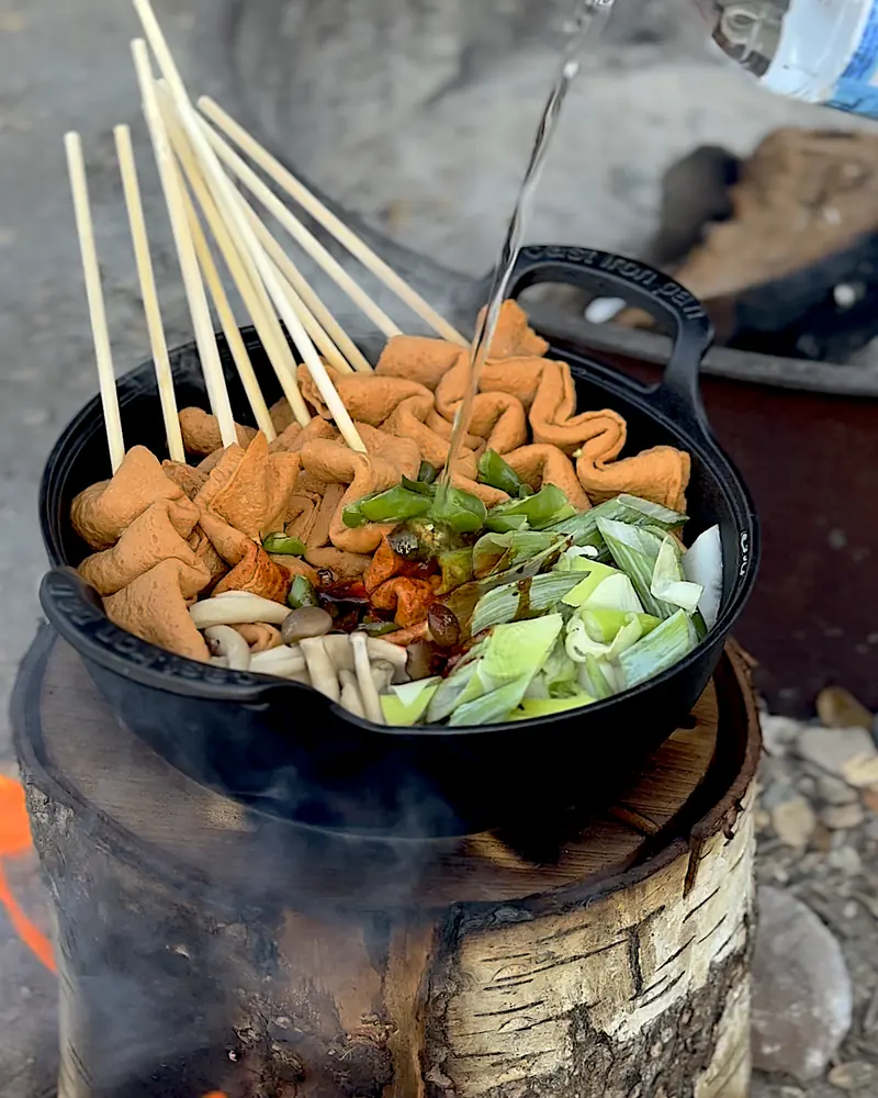 Cooking on a Fire Pit at Camp (6 Things We Made, From Lamb Chops to Pork Belly) Pouring water into the cast iron pot of fish cake soup on top of the Swedish fire log