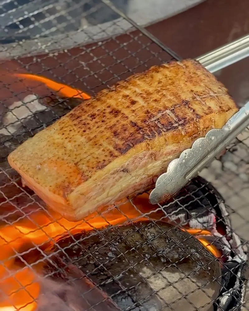 Cooking on a Fire Pit at Camp (6 Things We Made, From Lamb Chops to Pork Belly) Golden, crispy pork belly slab being turned with tongs on a wire rack over the fire